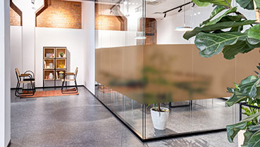 Modern office space with glass-walled meeting room featuring frosted film, plants, and exposed brick accents.